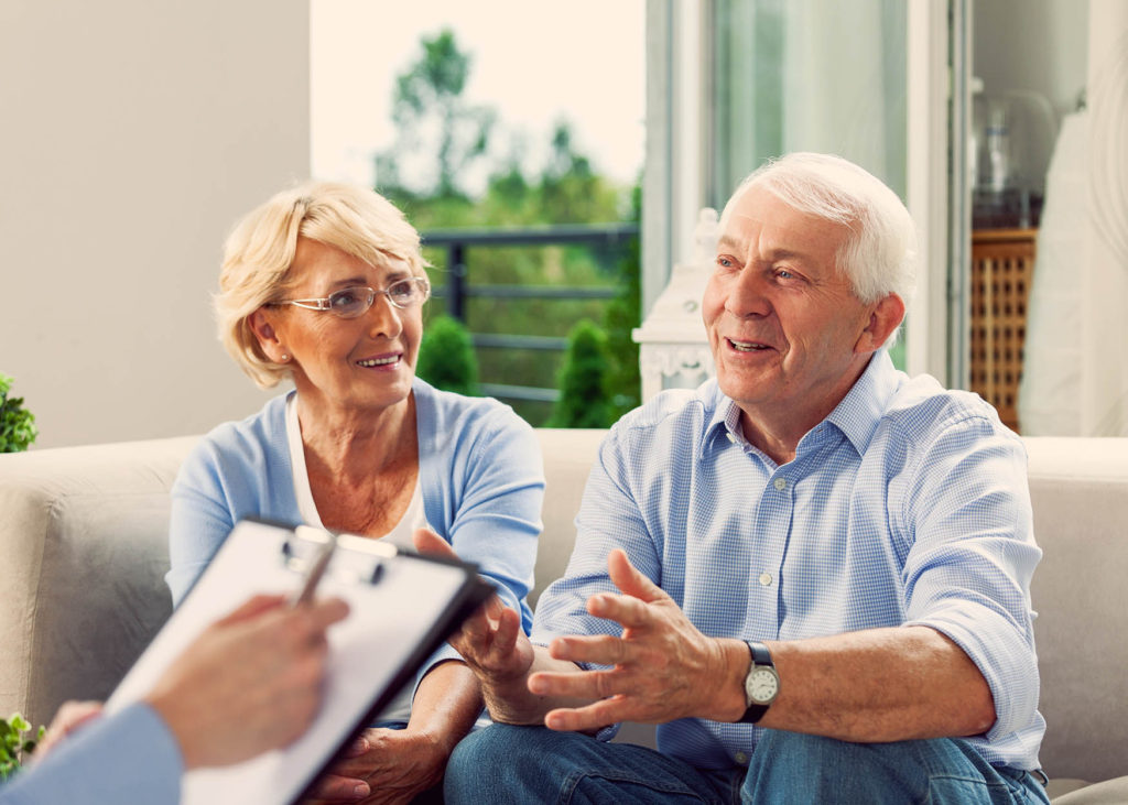 an elderly couple sit on a couch talking to a man holding a clipboard