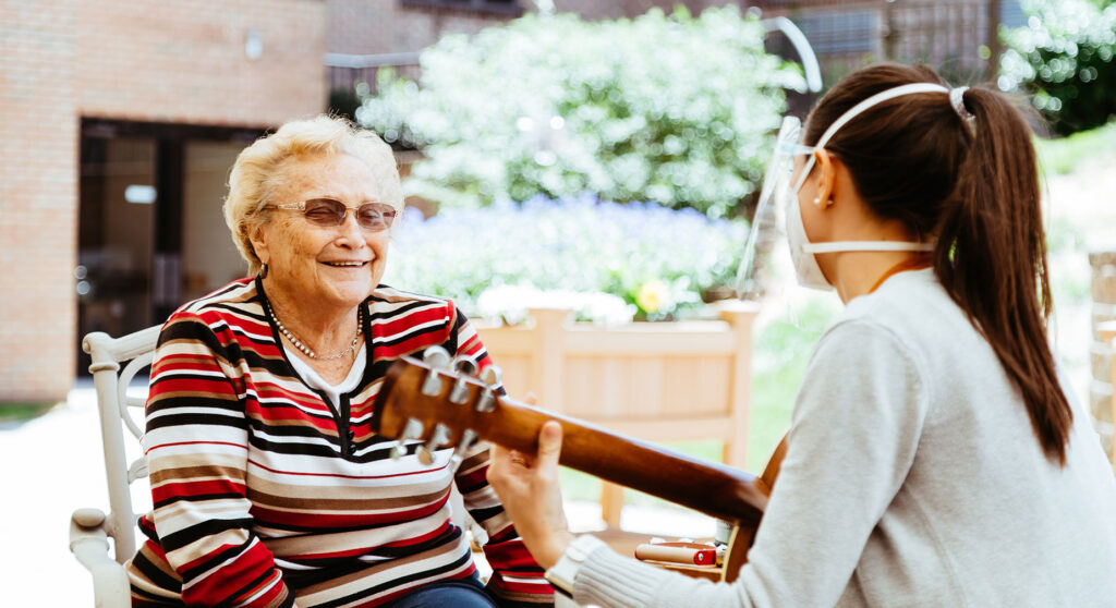 A young woman wearing a mask plays the acoustic guitar for a senior citizen