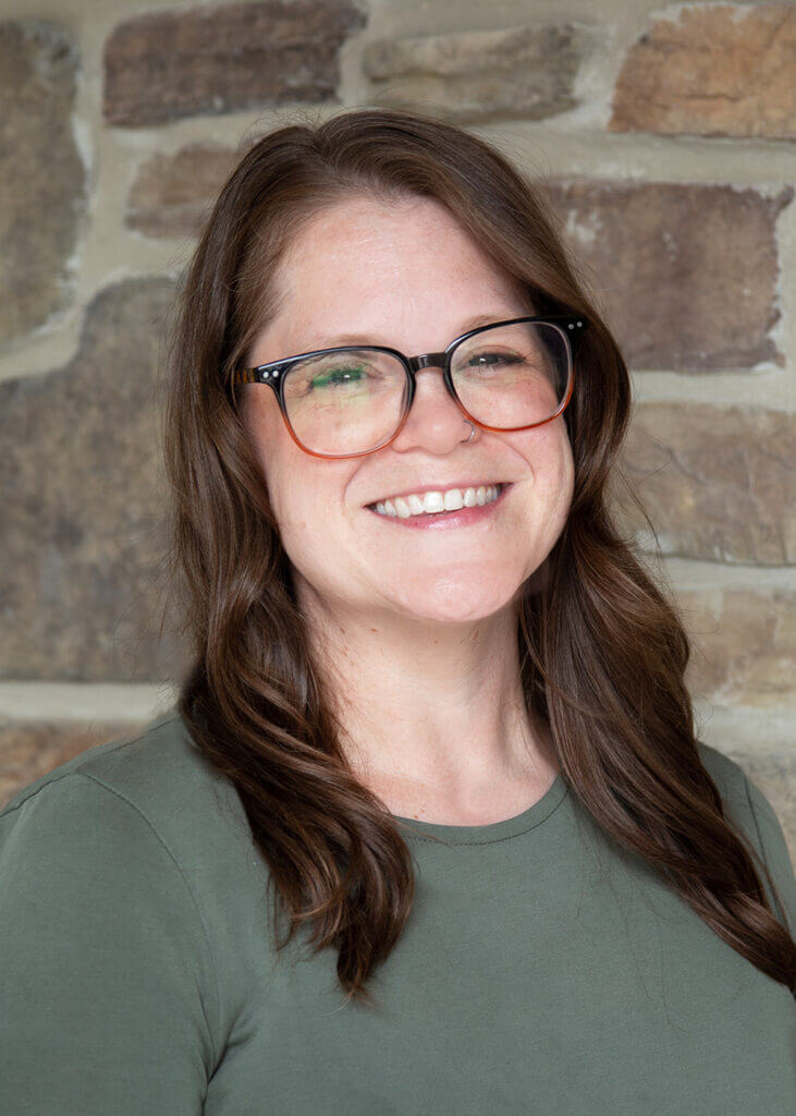 a woman wearing glasses and a green shirt smiles in front of a stone wall