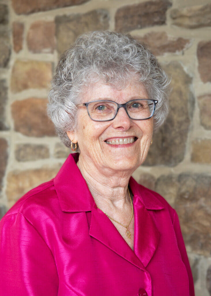 a woman wearing glasses and a pink shirt smiles in front of a stone wall