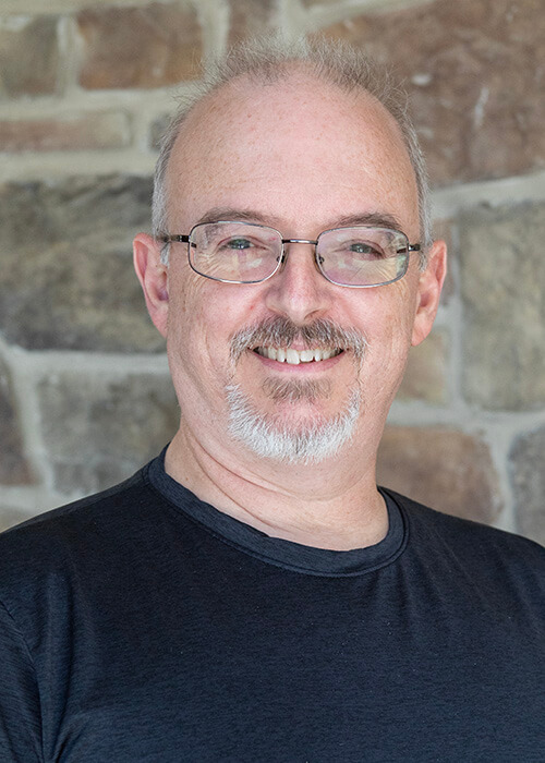 a man wearing glasses and a black shirt smiles in front of a stone wall