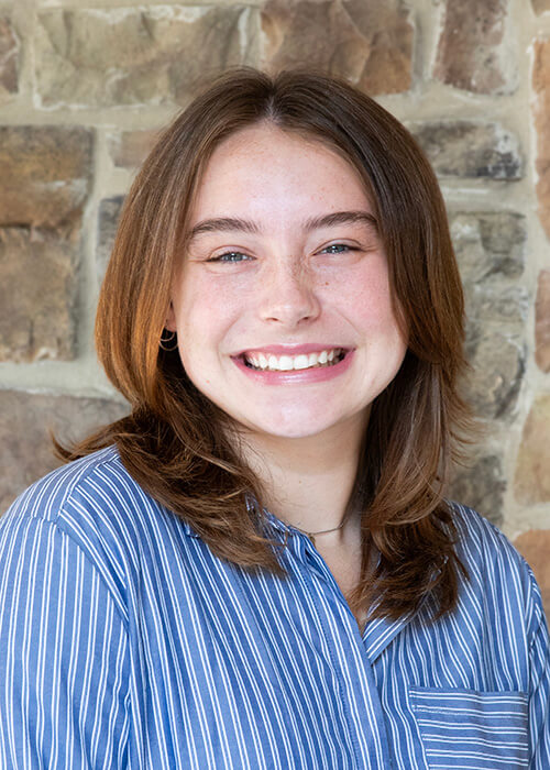 a woman in a blue striped shirt smiles in front of a stone wall